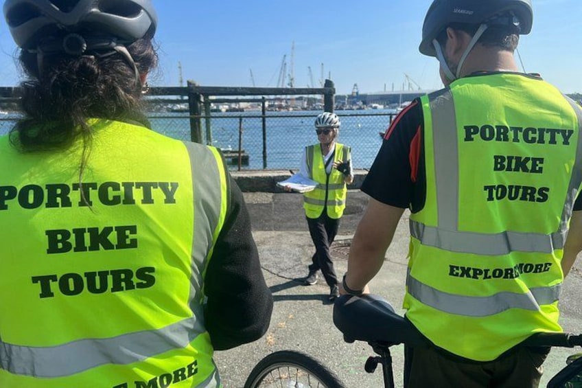 Two people wearing high-visibility vests with 'Portcity Bike Tours' on the back, standing by a waterfront with a bicycle. 2