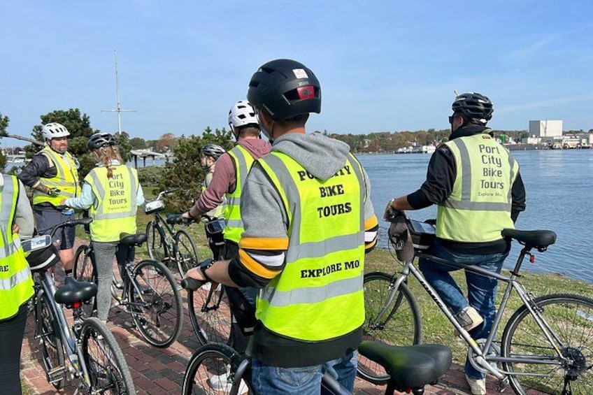 A group of people wearing reflective vests and helmets, standing by bicycles near a body of water. 2