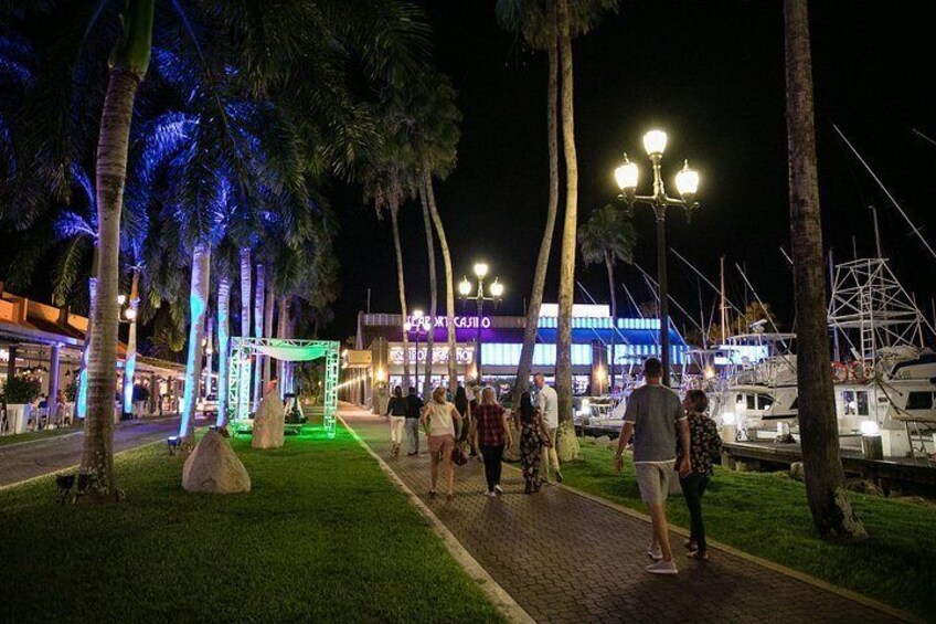 En promenadstig som är upplyst på natten, med palmer, en grönskimrande byggnad och båtar som ligger förtöjda i bakgrunden. 2