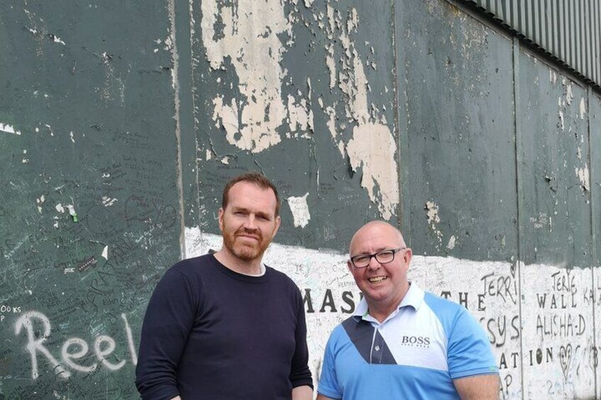 Dos hombres de pie frente a un muro con grafitis y pintura descascarada. 5