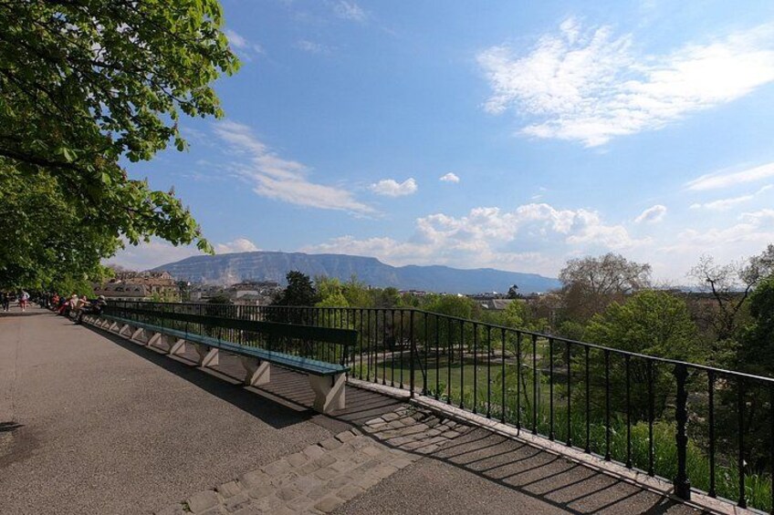 Un pont piétonnier doté d’une rambarde métallique, d’un banc vert et offrant une vue sur des montagnes et des arbres situés au loin. 3