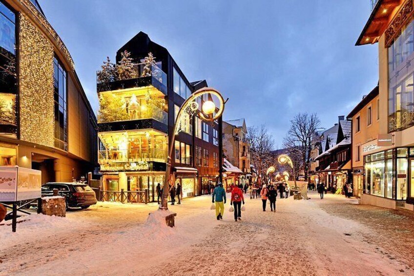 A snow-covered street with buildings, a street lamp, and people walking. 4