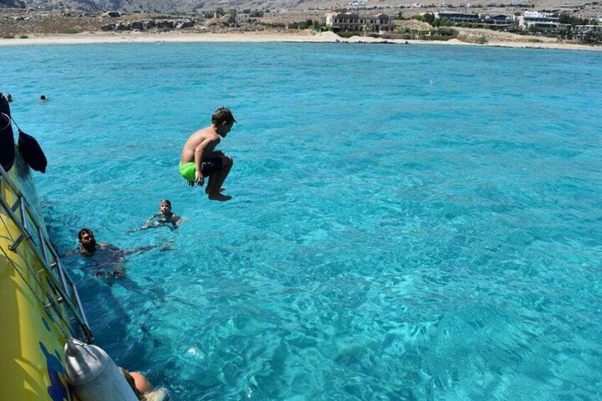 A person jumping into clear blue water from a boat, with buildings and a beach in the background. 1