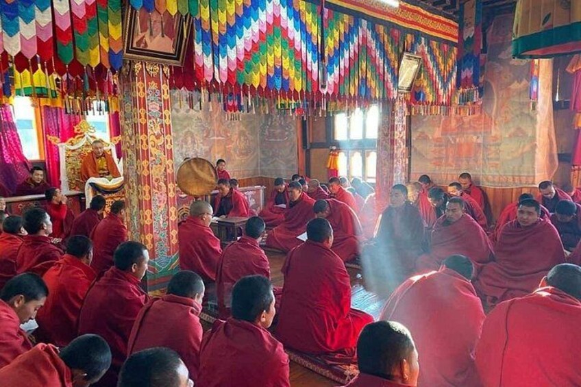 Monks in red robes seated in a temple with colorful decorations and a central figure on a throne. 4