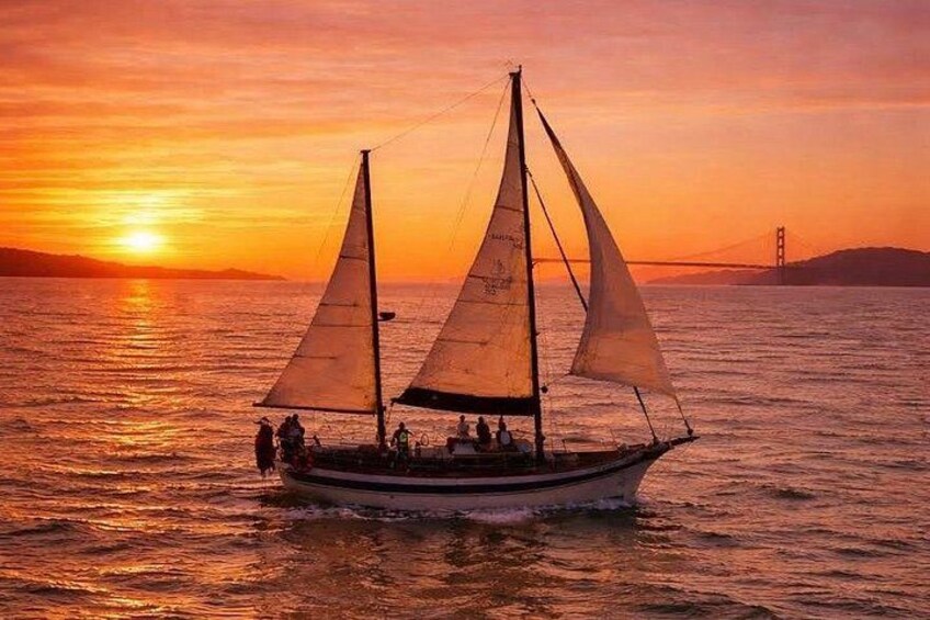 A sailboat with two sails on the water during sunset with a bridge in the background. 1