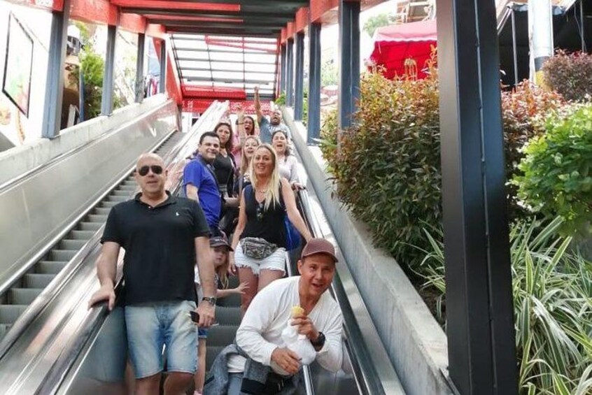 Un grupo de personas en una escalera mecánica al aire libre, con vegetación y un toldo rojo encima. 4