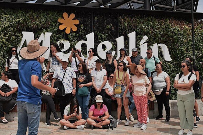 Un grupo de personas reunidas frente a un letrero grande con la palabra 'Medellín' y un símbolo de flor. 2