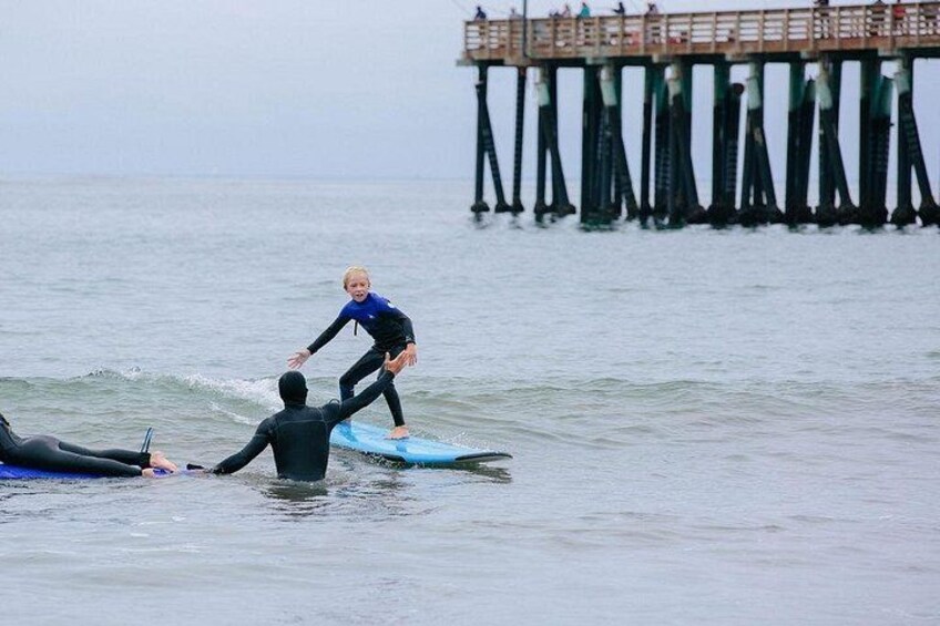 A person surfing on a blue board, another person in a wetsuit lying on a surfboard, and a pier in the background. 1