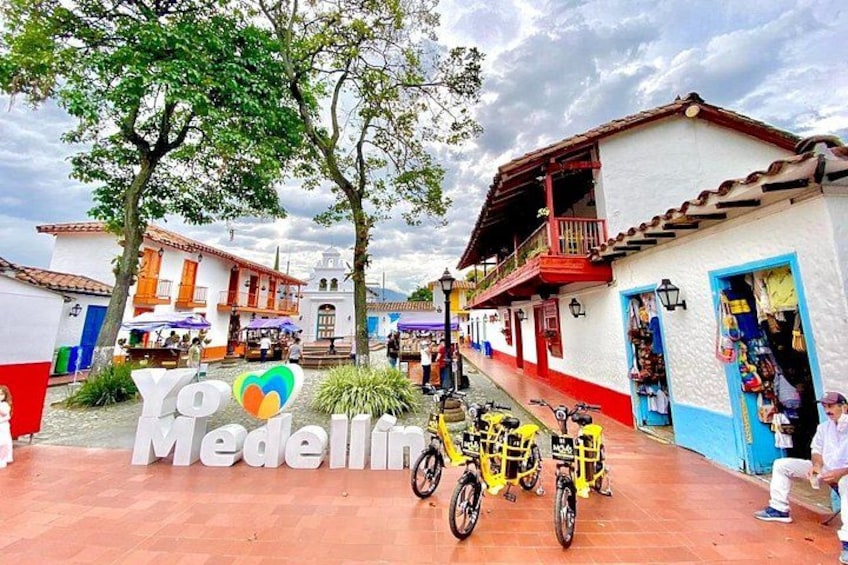 A street scene with traditional buildings, a sign reading 'Yo Medellín,' and yellow bicycles parked in front. 1