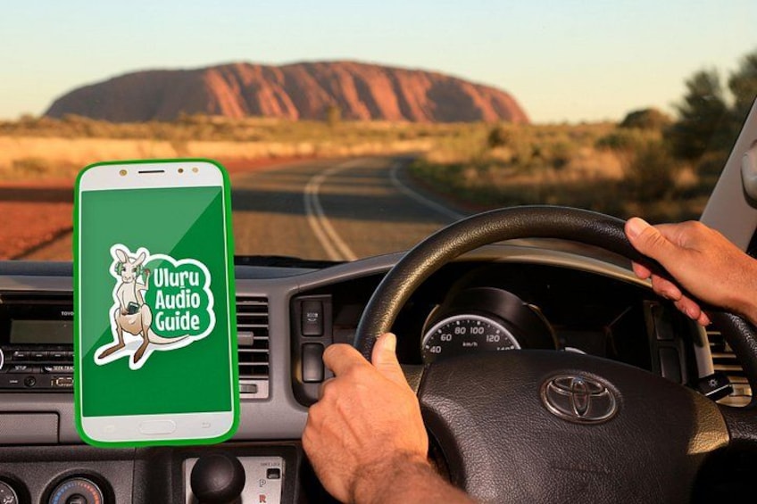 A smartphone displaying 'Uluru Audio Guide' on a dashboard, with a steering wheel and a view of Uluru in the background. 1