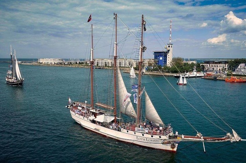 A sailing ship named 'Loth Loris' docked at a harbor with other boats, a lighthouse, and buildings in the background. 3