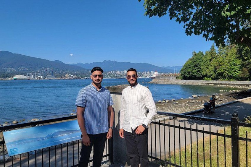 Two men standing by a waterfront with a cityscape and mountains in the background. 2