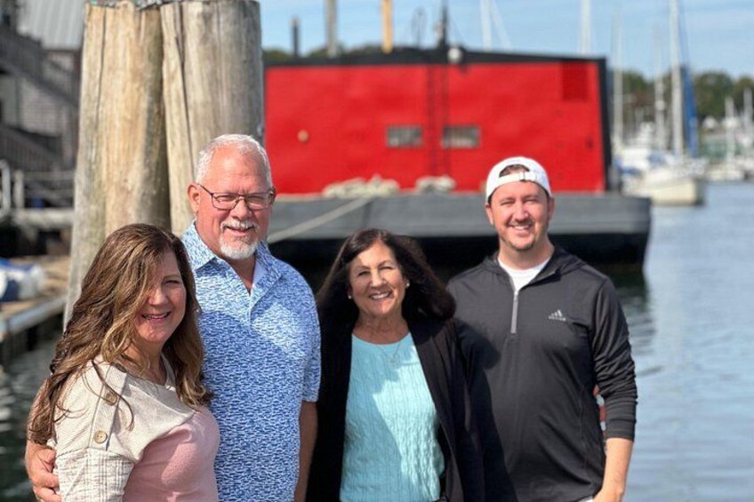 A group of people standing by a waterfront with a red building and boats in the background. 5