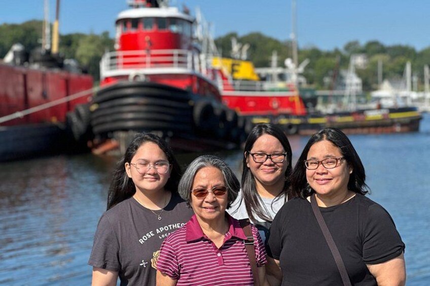 A group of people standing in front of a large red and black ship docked at a harbor with other boats in the background. 3