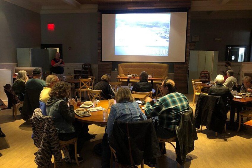 A group of people seated at tables in a room with a presentation screen at the front. 2