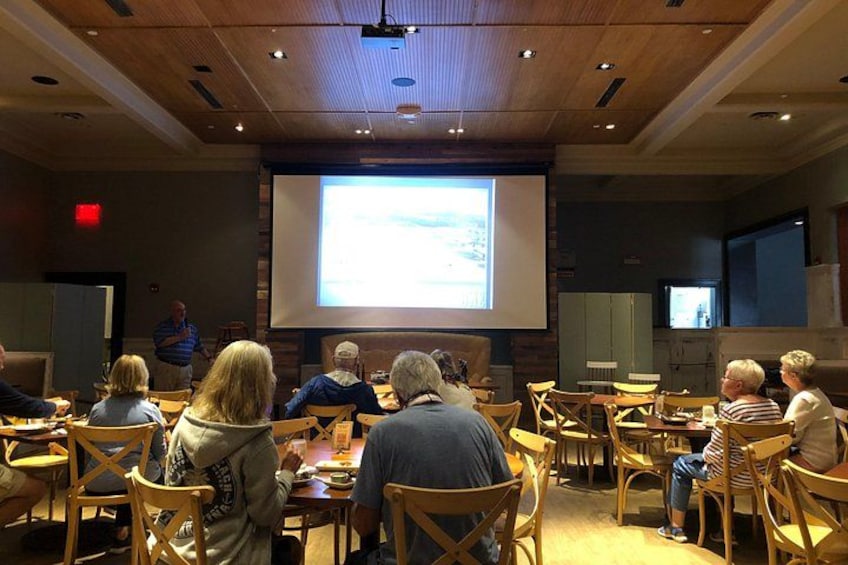 A group of people seated at round tables in a conference room with a projector screen in the background. 3