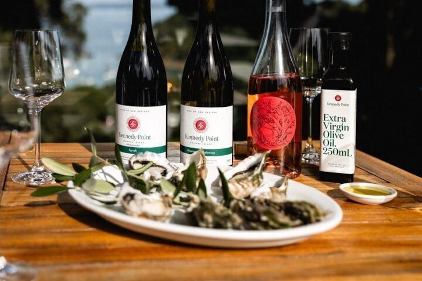 A wooden table with two bottles of Kennedy Point wine, a bottle of Extra Virgin Olive Oil, and a plate of oysters. 1