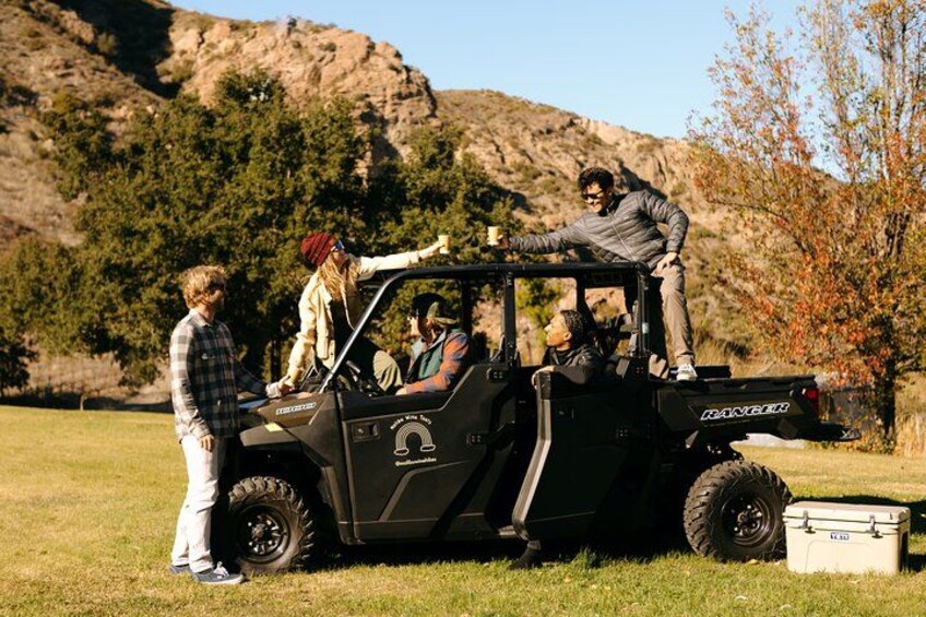 A black Maxxair vehicle with four people, one standing on the roof, parked on grass with a rocky hill in the background. 1
