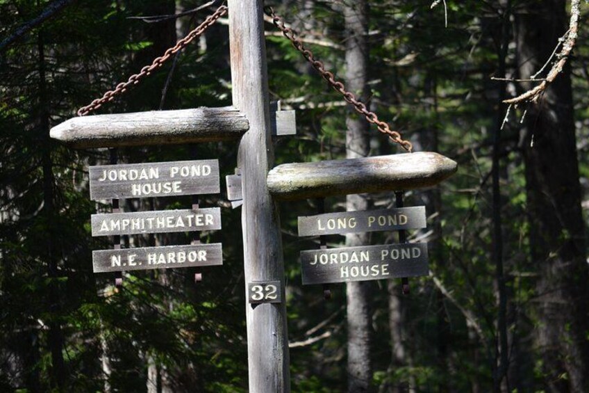 A wooden signpost with multiple directional signs pointing to locations such as Jordan Pond House, Amphitheater, N.E. Harbor, and Long Pond. 2