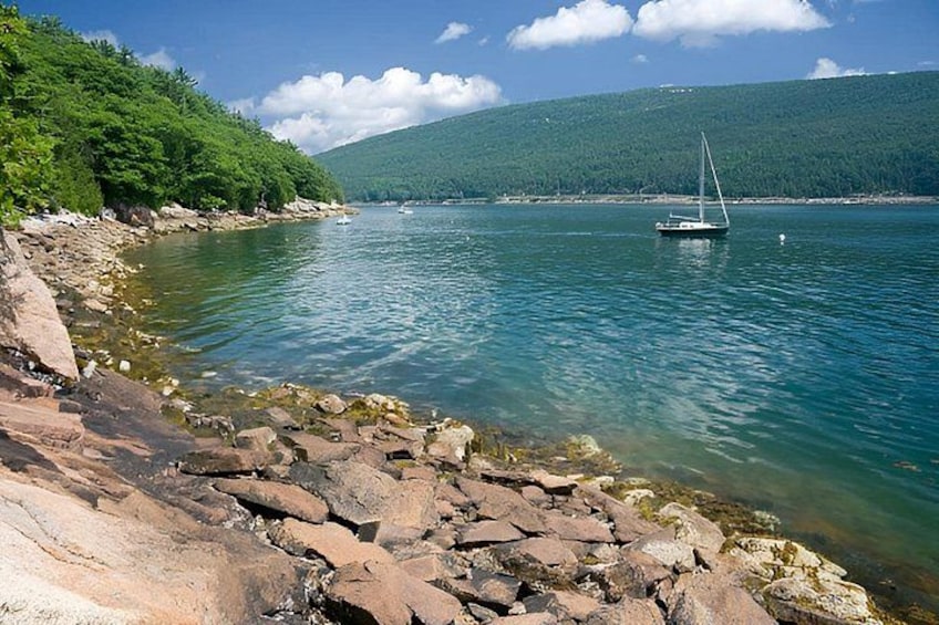 A calm lake with a sailboat, rocky shoreline, and a forested hill in the background. 2