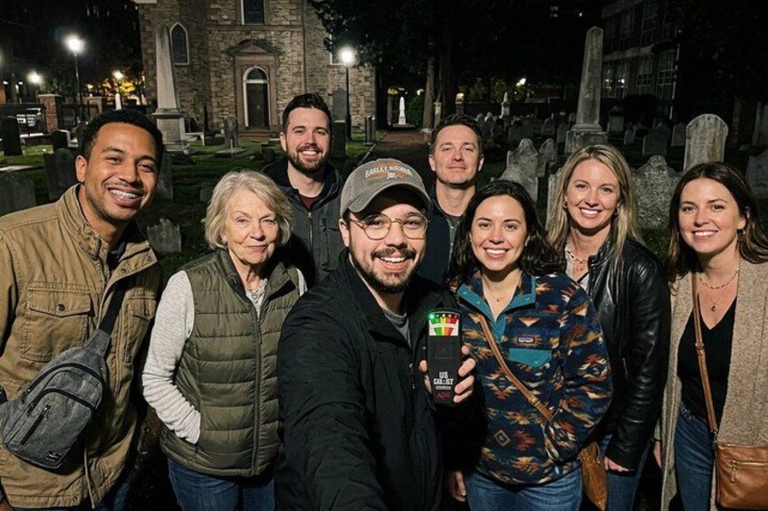 A group of people posing for a photo in a cemetery with tombstones and a building in the background. 1