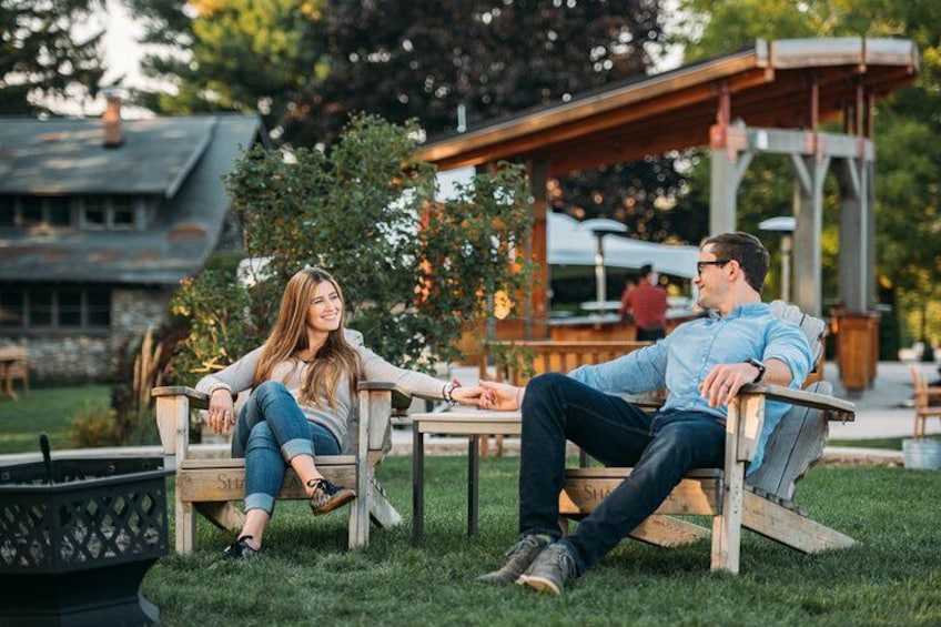 Twee personen zitten op houten stoelen bij een terras met een tuin en een gebouw op de achtergrond. 2