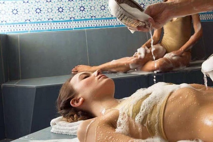 Eine Person erhält eine Gesichtsbehandlung mit einem Pinsel in einem Badezimmer mit blau-weißen Fliesen. 1