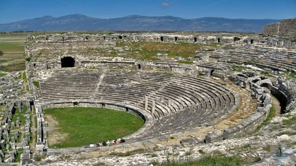 Un ancien amphithéâtre avec des gradins en pierre et une scène centrale, entouré de ruines et dominé par des montagnes en arrière-plan. 5