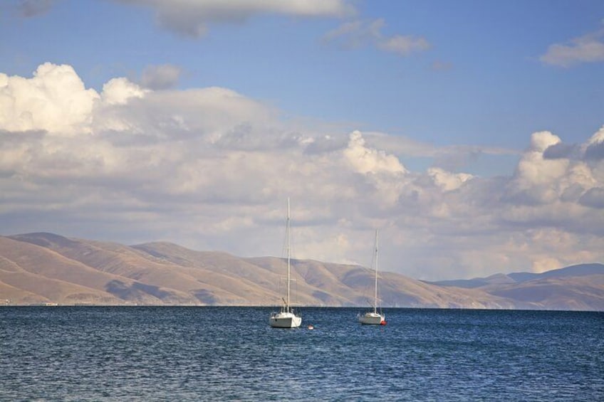 A calm sea with two sailboats anchored, surrounded by rolling hills under a partly cloudy sky. 3