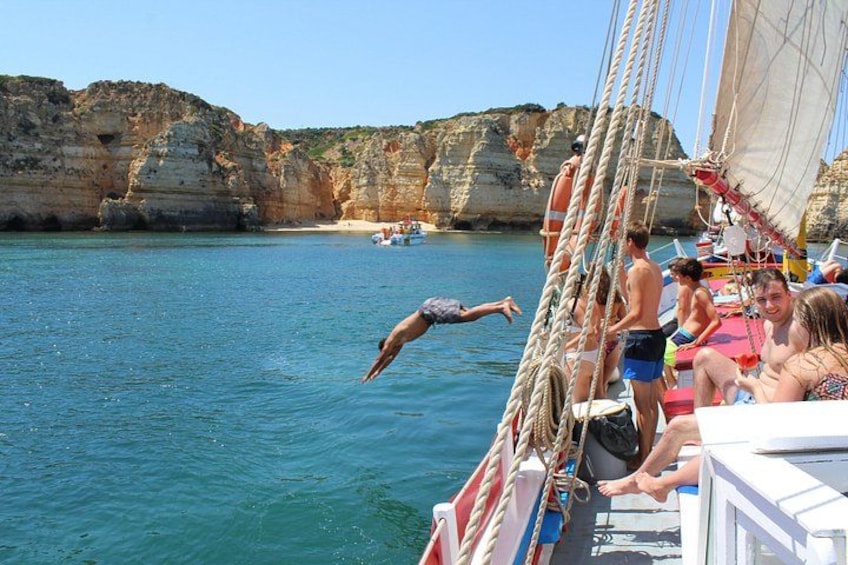 A sailboat with people on board, a rocky coastline, and clear blue water. 1