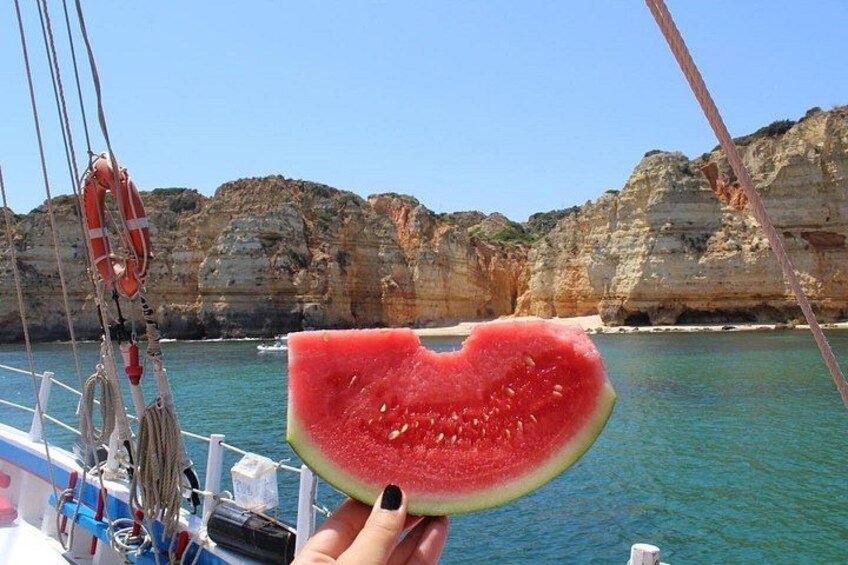 A slice of watermelon held by a person with black nail polish, with a rocky coastline and clear blue water in the background. 3