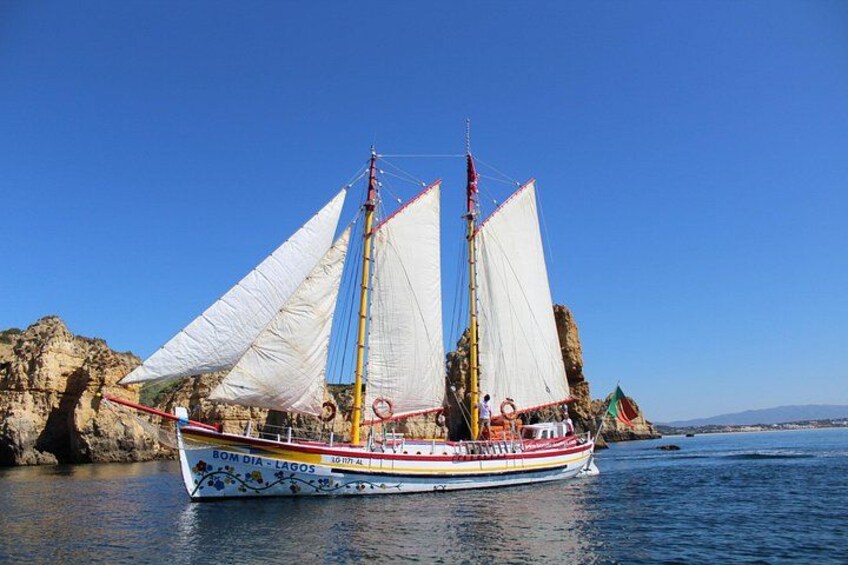 A traditional sailing boat with white sails and a colorful hull, anchored near rocky cliffs. 2