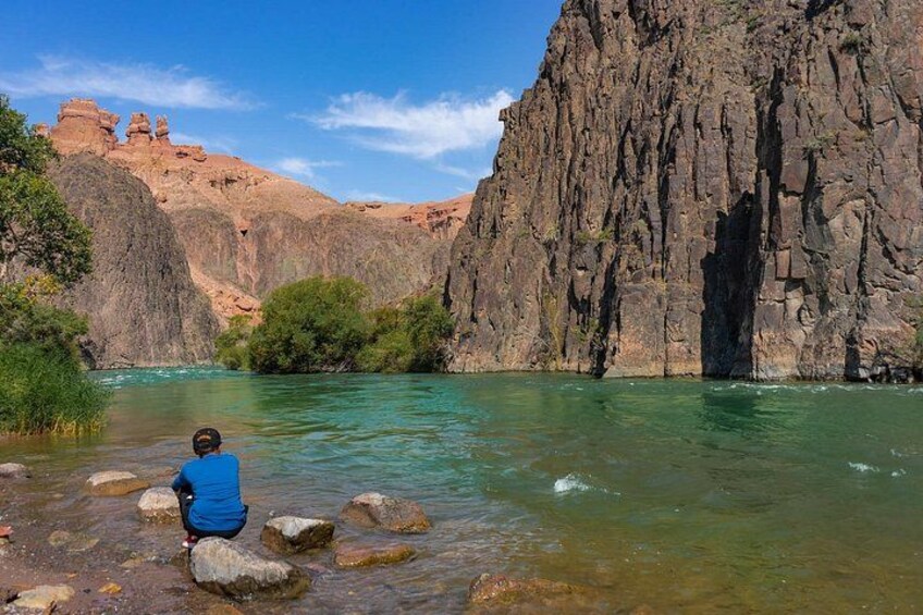 A person sitting by a clear river with rocky cliffs in the background. 2
