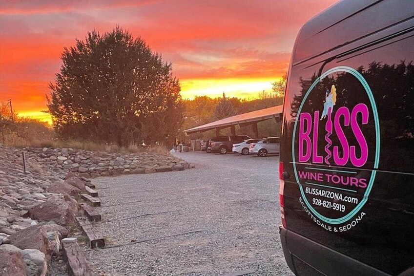 A black van with 'BLISS WINE TOURS' branding, parked in a gravel area with a sunset in the background. 3