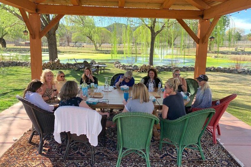 A group of people sitting at an outdoor table under a wooden pergola, with a grassy area and trees in the background. 4