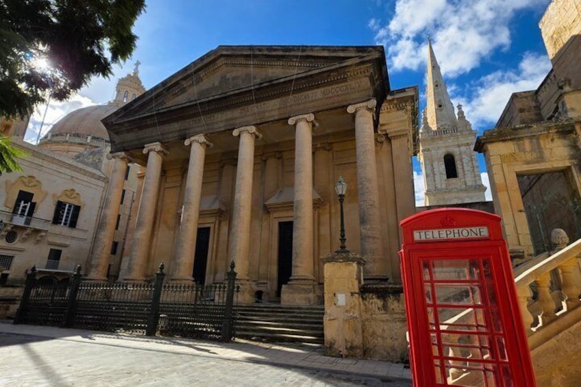 A classical building with columns, a red telephone booth, and a church spire in the background. 5
