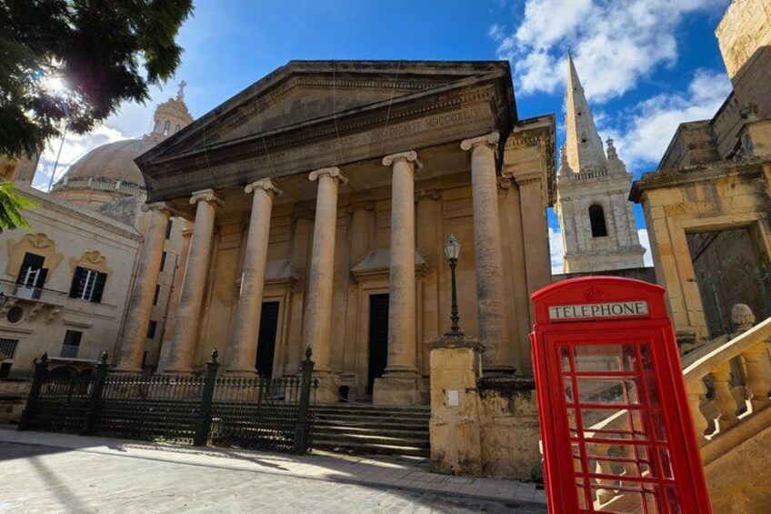 A classical building with columns, a red telephone booth, and a church spire in the background. 2