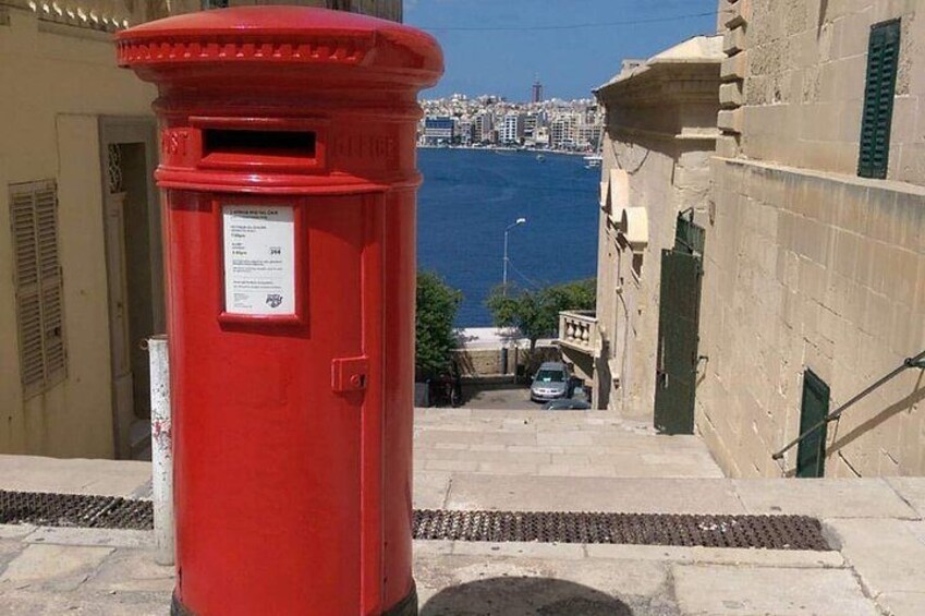 A red post box on a stone staircase with a view of the sea and buildings in the background. 4