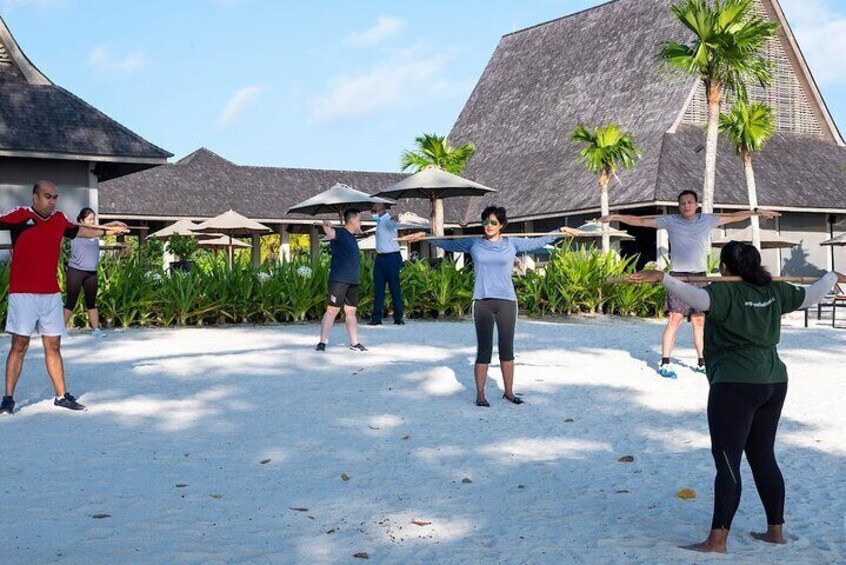 A group of people exercising on a sandy beach with palm trees and resort buildings in the background. 1
