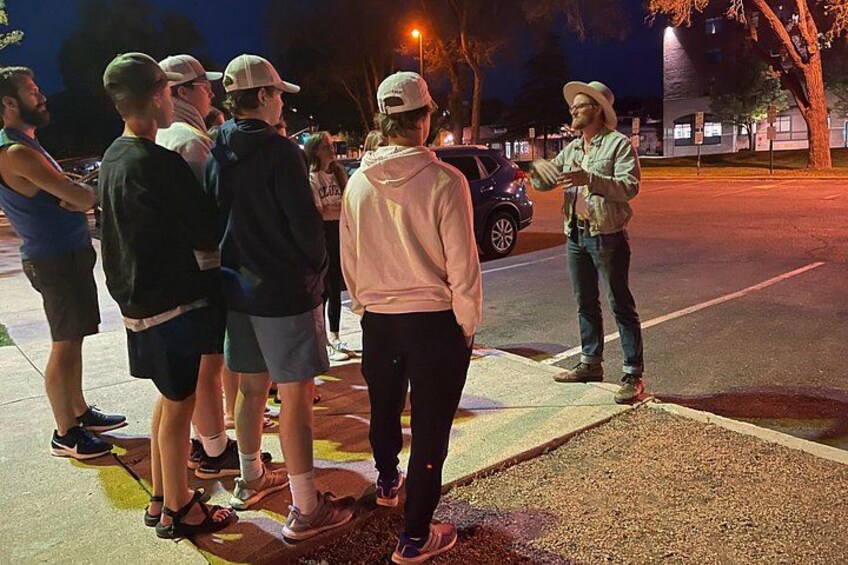 Un grupo de personas de pie en una acera de noche, con una farola y edificios al fondo. 2
