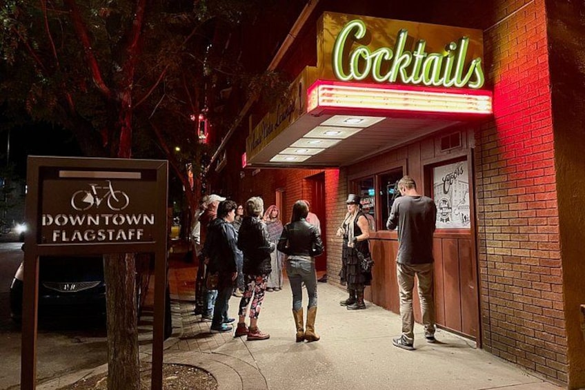 Ein leuchtendes Neon-Schild mit der Aufschrift 'Cocktails', ein Schild mit 'Downtown Flagstaff' und eine Gruppe von Personen, die auf dem Bürgersteig stehen. 2