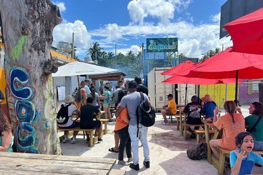 A crowded outdoor dining area with people sitting at wooden tables under red umbrellas, a tree with graffiti, and a sign for 'mojitoal'. 1