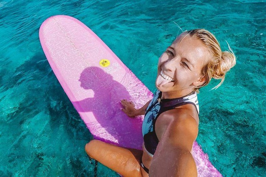 Une personne assise sur une planche de surf rose, dans une eau d’un bleu limpide. 2