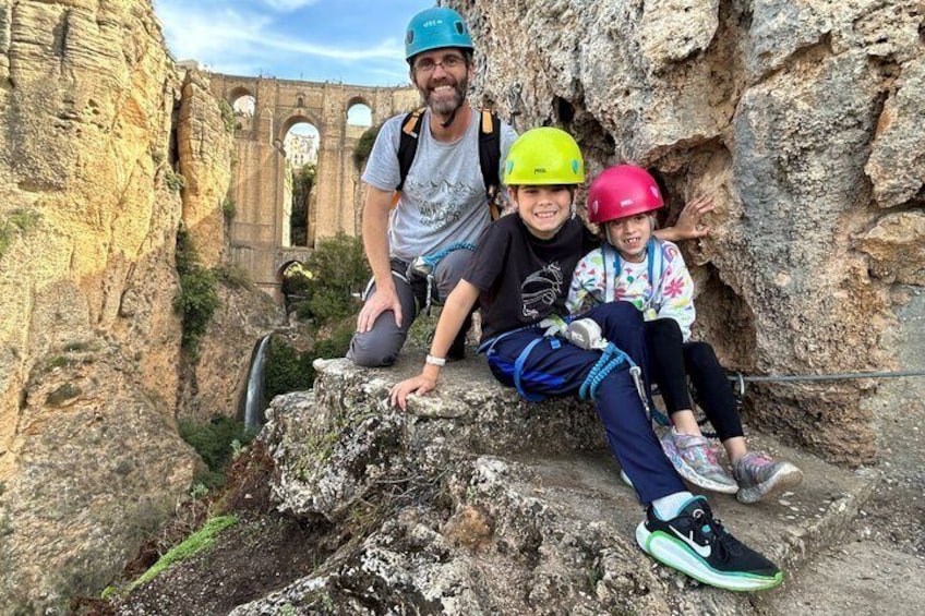 Three people sitting on a rock ledge with a historical bridge in the background. 3