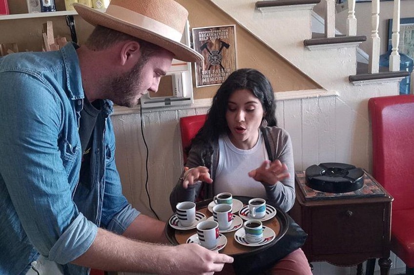 Zwei Personen sitzen an einem Tisch mit Tassen und Untertassen, einem Hut und einer kleinen Tafel mit einem dekorativen Gegenstand. 4