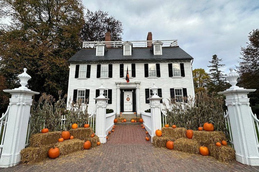 A white house with black shutters, a front porch, and hay bales with pumpkins. 2