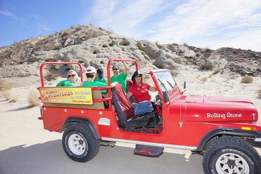 Un jeep rojo con la leyenda 'Rolling Stone' en la carrocería, estacionado en una carretera con un paisaje desértico pedregoso al fondo. 3