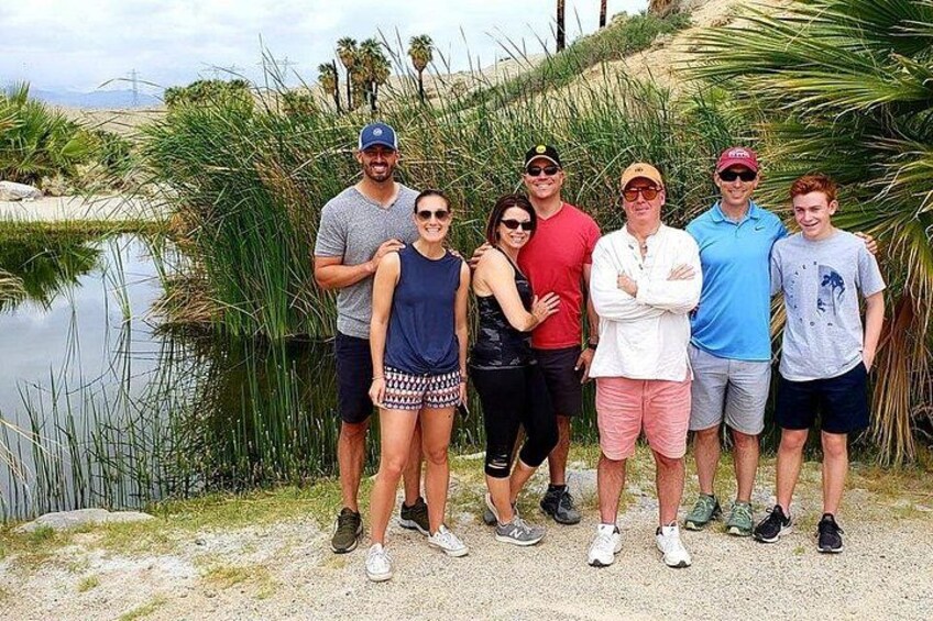 Un grupo de personas de pie junto a un cuerpo de agua, con palmeras y una colina al fondo. 2
