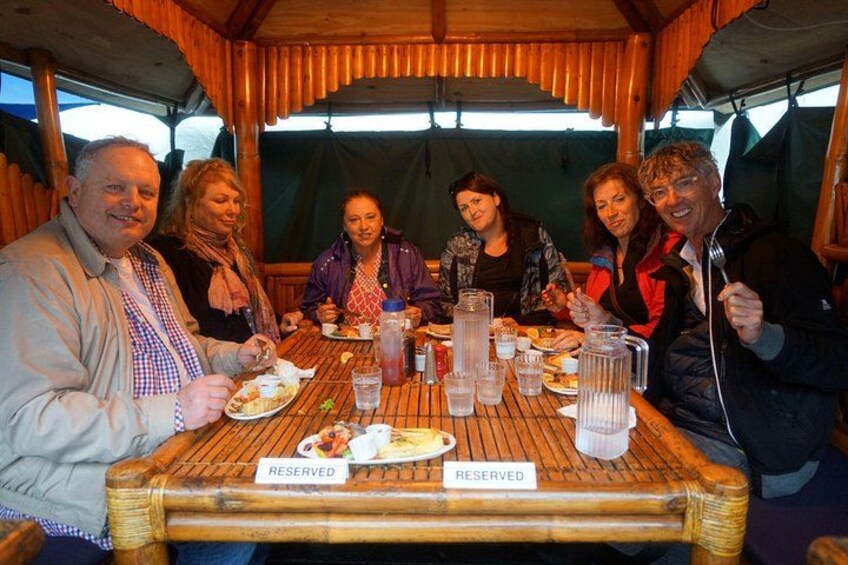 A group of people dining at a bamboo table with a 'RESERVED' sign, under a wooden canopy. 2