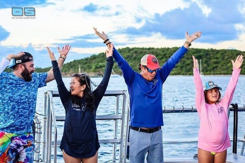 A group of people on a boat with their arms raised, a body of water, and a forested shoreline in the background. 2